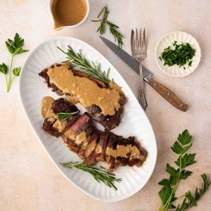 A sliced steak topped with creamy sauce is served on a white platter with rosemary sprigs, accompanied by a small bowl of chopped herbs and utensils.