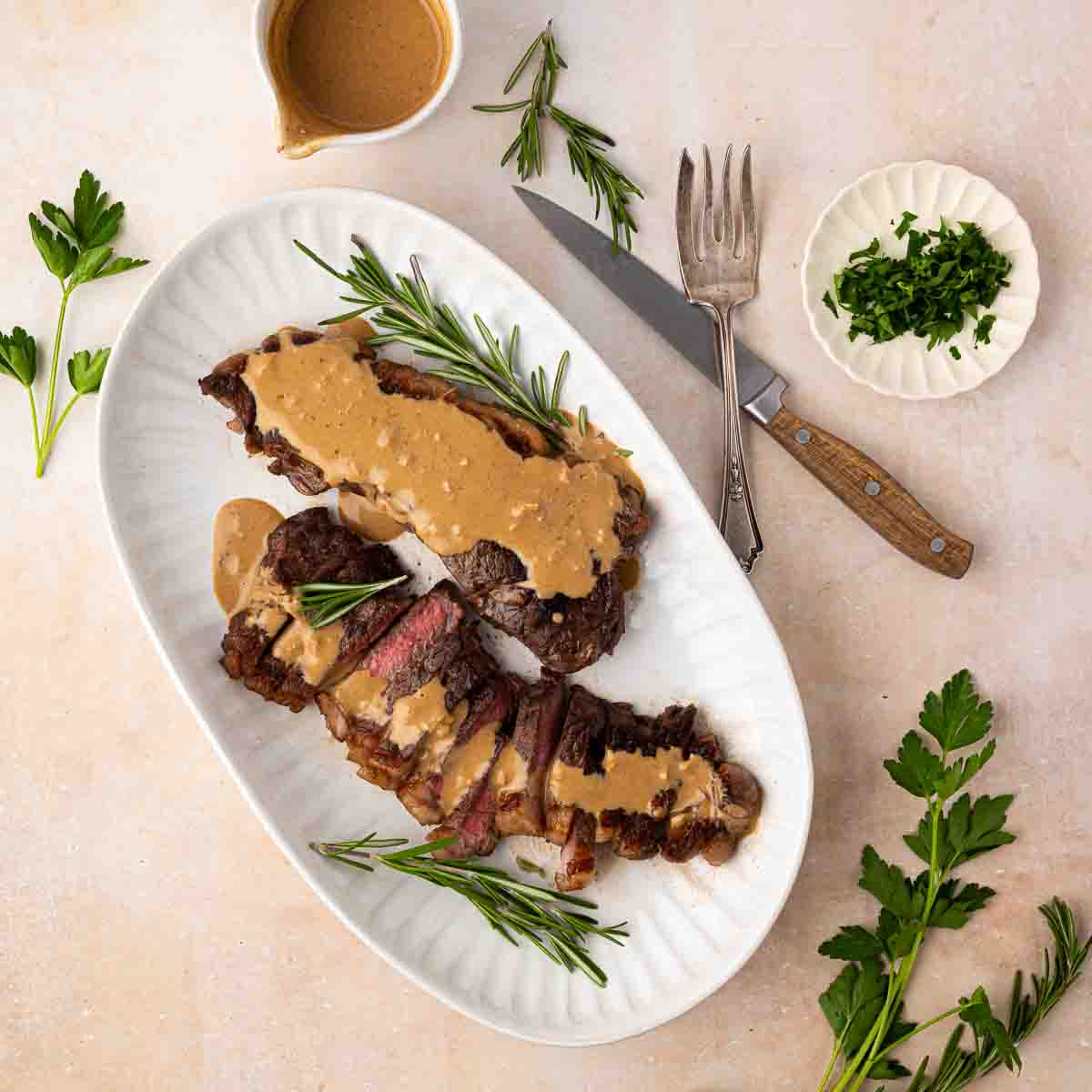 A sliced steak topped with creamy sauce is served on a white platter with rosemary sprigs, accompanied by a small bowl of chopped herbs and utensils.