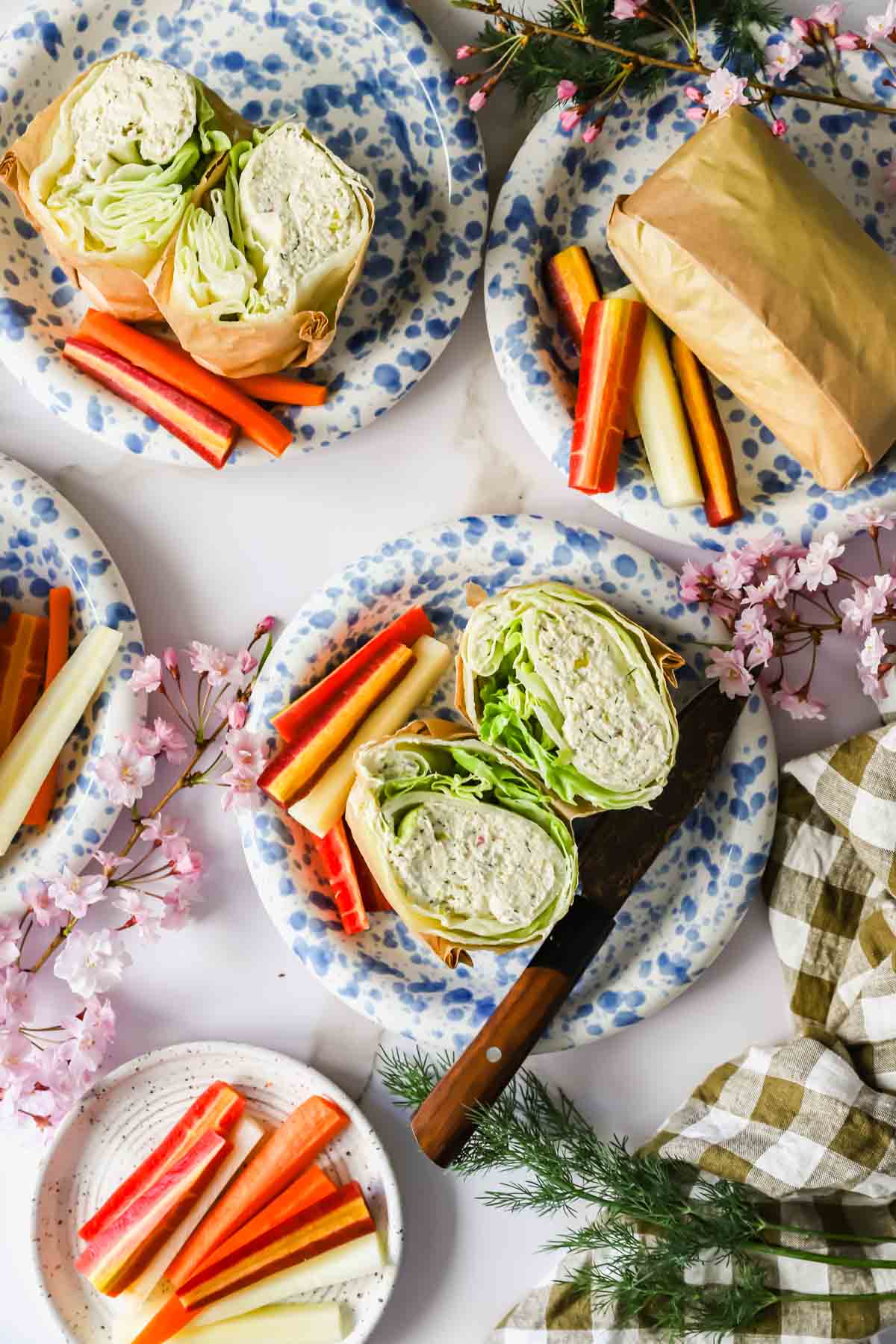 Four plates with lettuce wraps cut in half and colorful vegetable sticks, arranged on a white surface with flowers and a knife nearby.