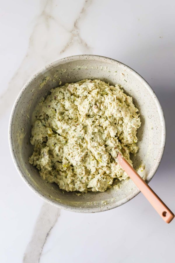 A mixing bowl filled with a creamy, textured mixture and a pink spatula resting inside on a white marble surface.