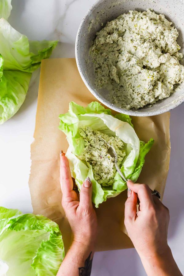 Hands scooping a mixture from a bowl onto a large lettuce leaf on parchment paper, preparing lettuce wraps.