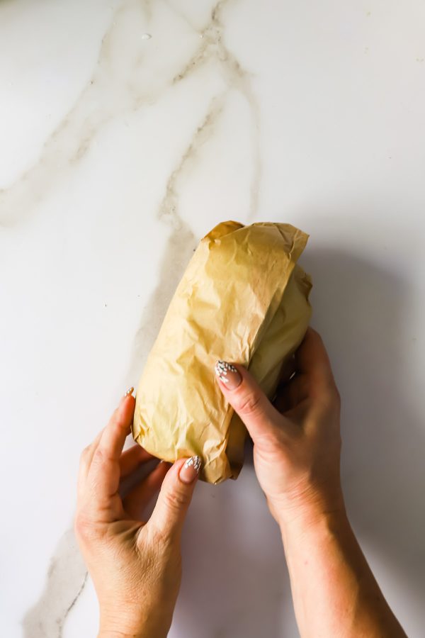 Two hands are holding a sandwich wrapped in brown paper on a white marble surface.