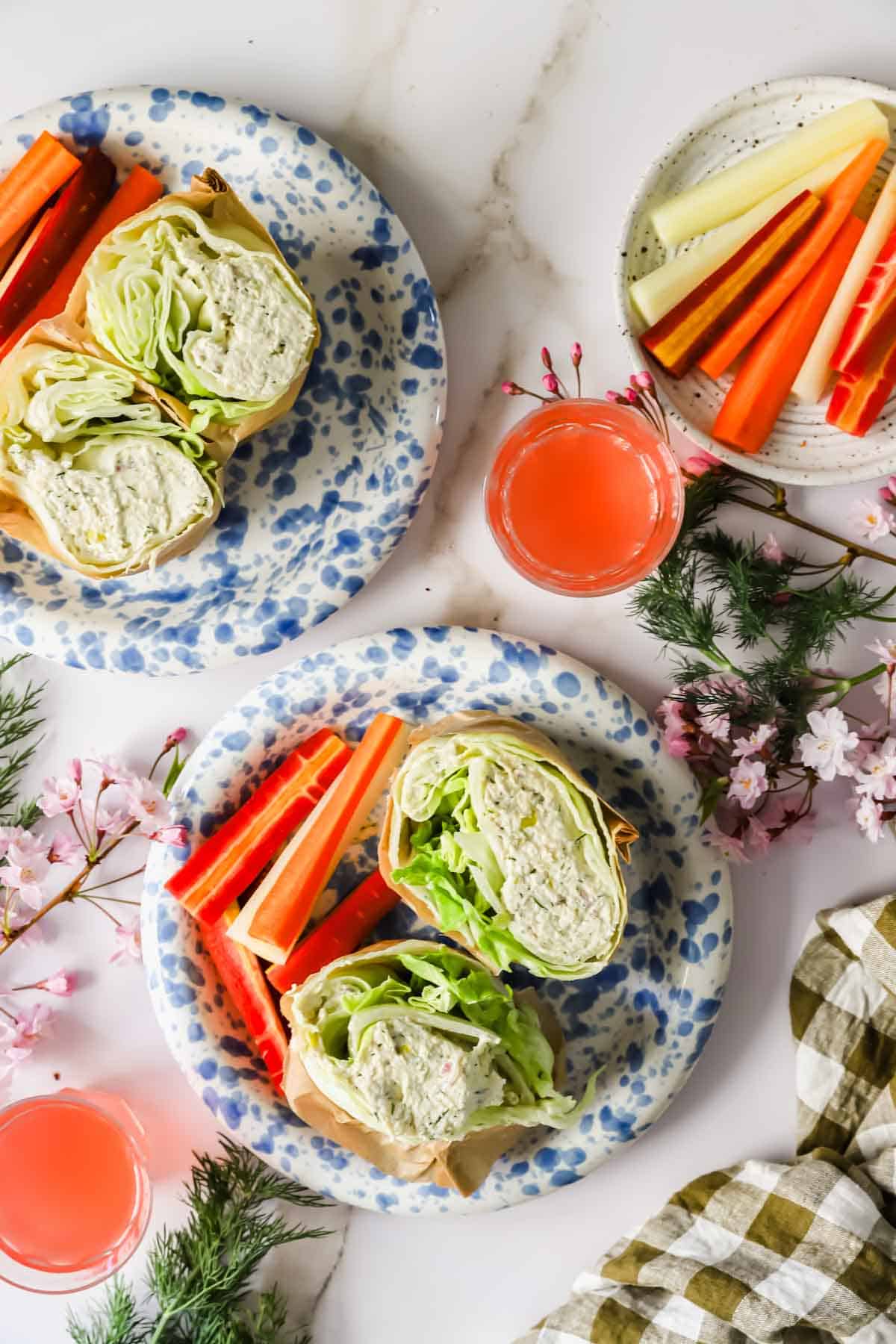 Two plates with chicken salad wraps, carrot and celery sticks, and two glasses of pink juice on a white surface with floral and greenery decorations.