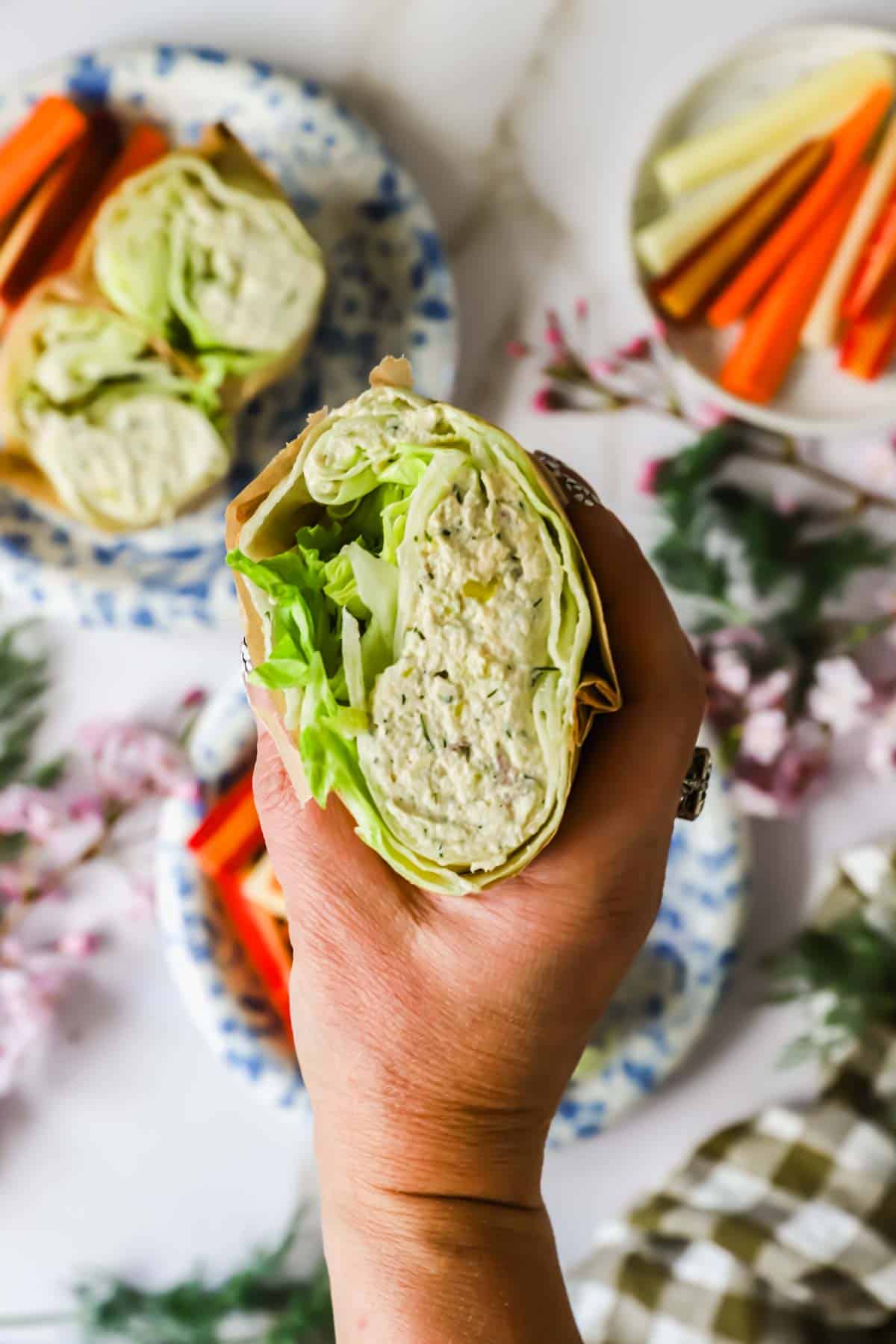 A hand holds a wrap filled with lettuce and a creamy mixture, with plates of sliced carrots and celery in the background.