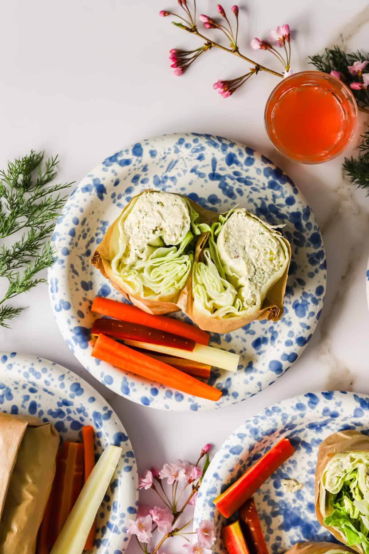 A plate with a lettuce wrap cut in half and carrot and celery sticks, surrounded by floral decorations and a glass of pink beverage.