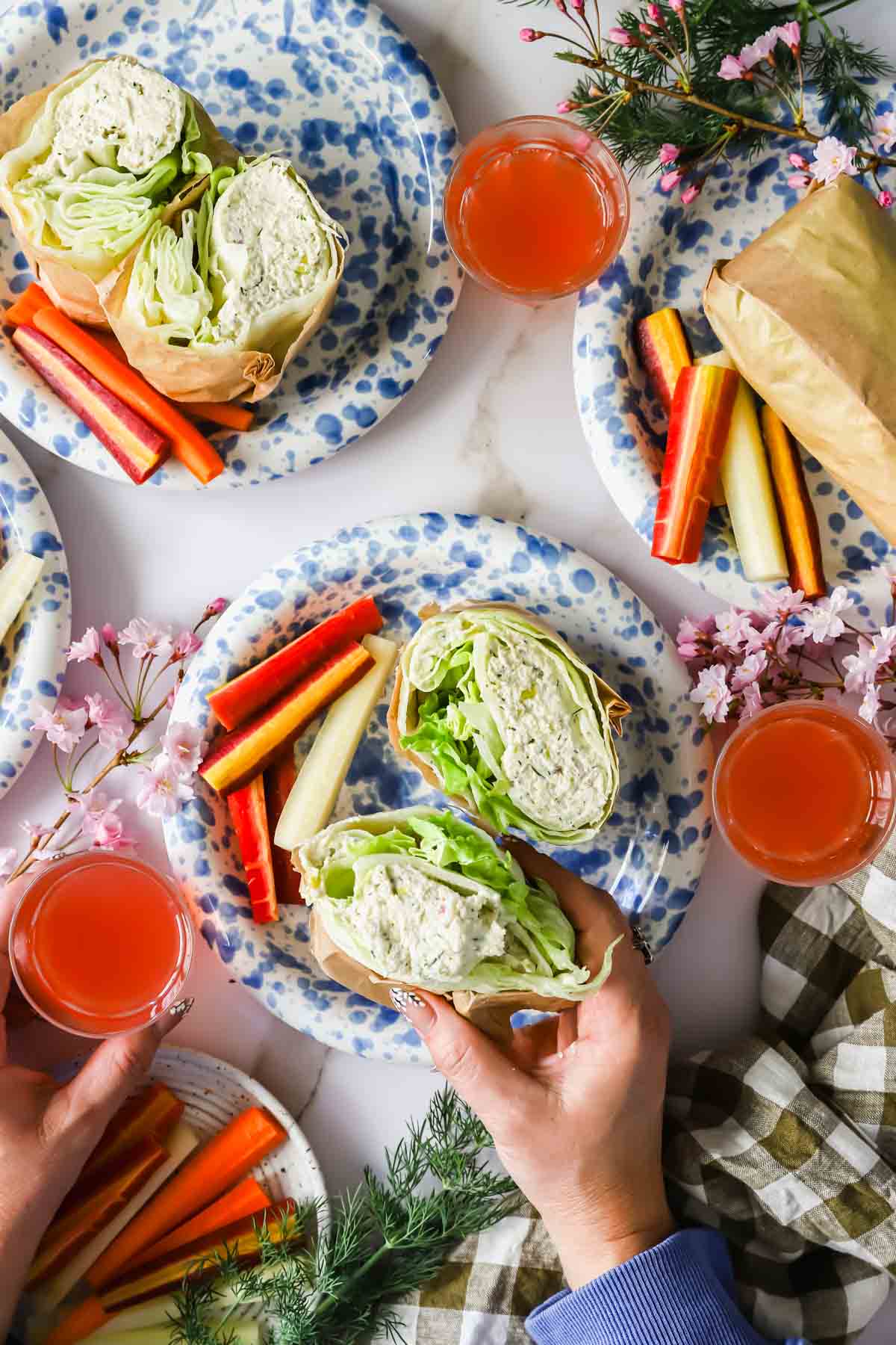 Four plates with lettuce wraps filled with chicken salad, assorted carrot sticks, pink drinks, and flower decorations on a table; hands hold one wrap.