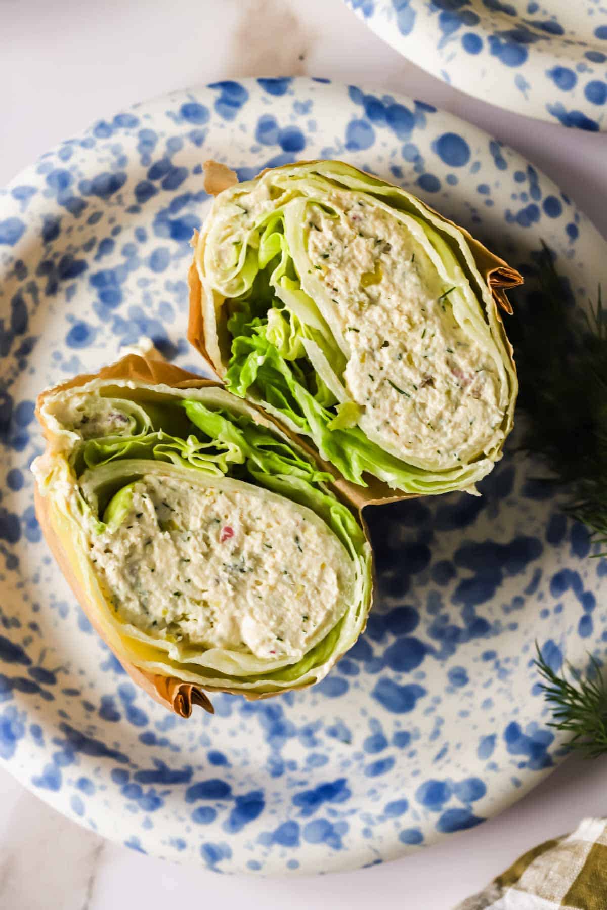 A wrap filled with chicken salad and lettuce, cut in half and placed on a white plate with blue speckles.