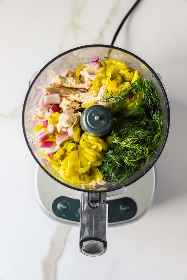 A food processor containing shredded chicken, banana peppers, red onion, and fresh dill on a white countertop.
