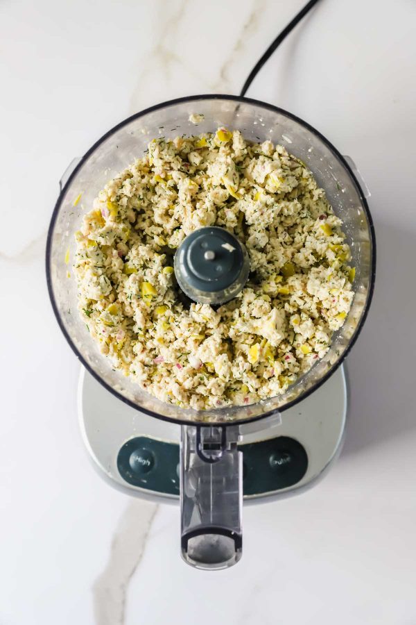 A food processor filled with finely chopped ingredients, including herbs and vegetables, sits on a white countertop.