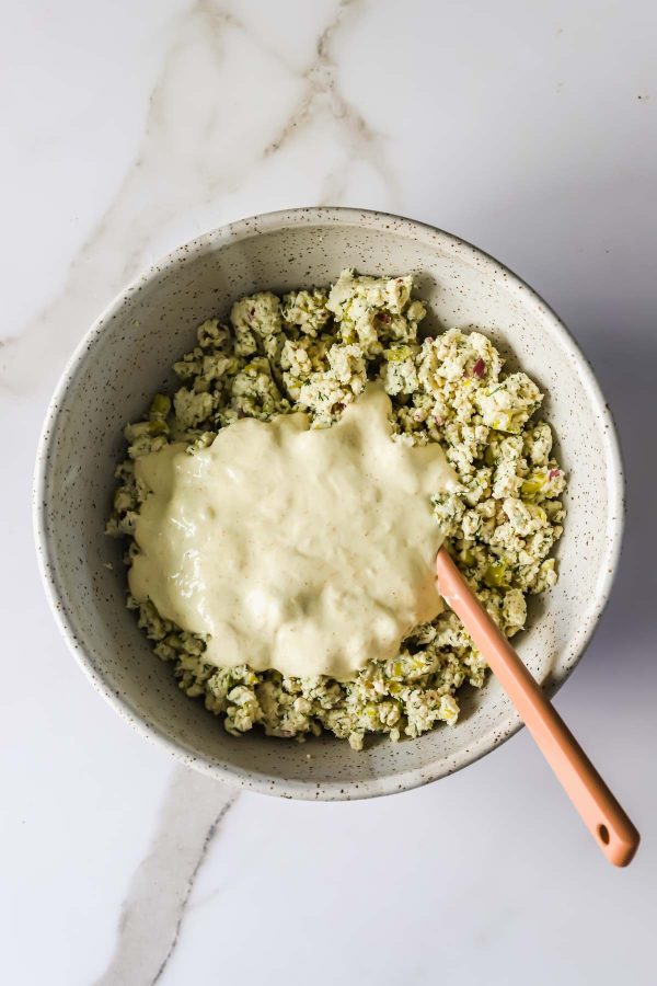 A large bowl containing chicken mixed with herbs, topped with a creamy white sauce, and a peach-colored spatula resting inside.