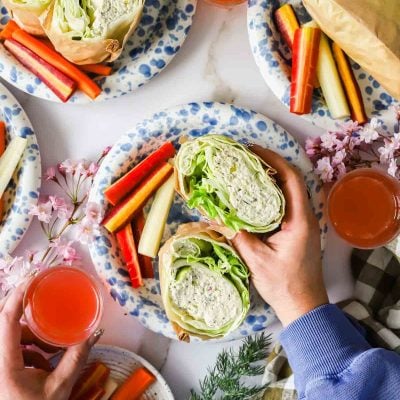 Hands holding a lettuce and chicken salad wrap on a plate with carrot and cucumber sticks, surrounded by drinks and floral decorations.