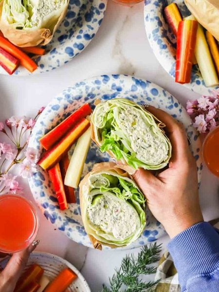 Hands holding a lettuce and chicken salad wrap on a plate with carrot and cucumber sticks, surrounded by drinks and floral decorations.
