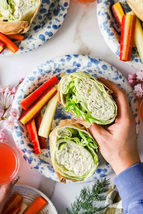 Hands holding a lettuce and chicken salad wrap on a plate with carrot and cucumber sticks, surrounded by drinks and floral decorations.