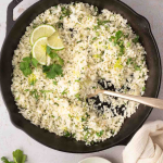 A skillet filled with cilantro lime cauliflower rice, garnished with lime slices and herbs, with a spoon, fresh cilantro, and lime wedges on the side.