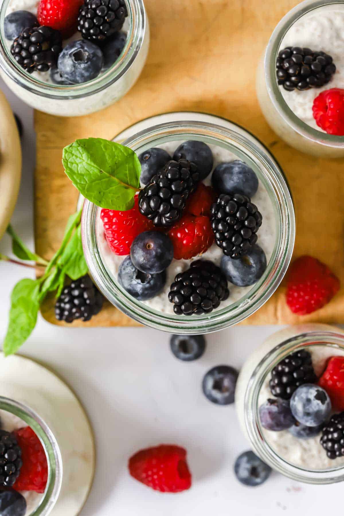 Jars of chia pudding topped with blackberries, blueberries, raspberries, and a sprig of mint are arranged on a wooden board.