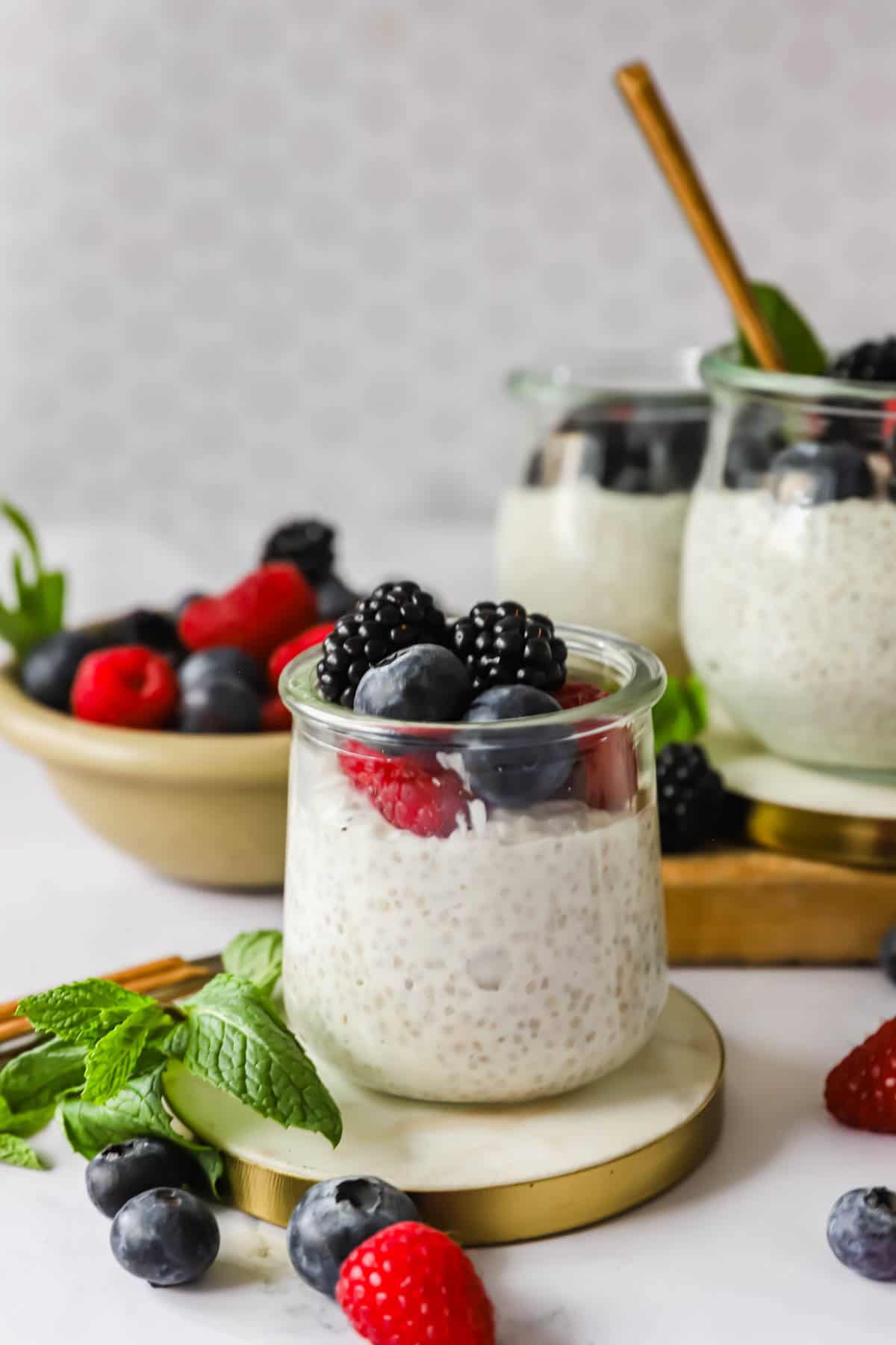 Glass jars filled with chia pudding topped with fresh blackberries, blueberries, and raspberries, garnished with mint leaves; extra berries and mint are arranged nearby.
