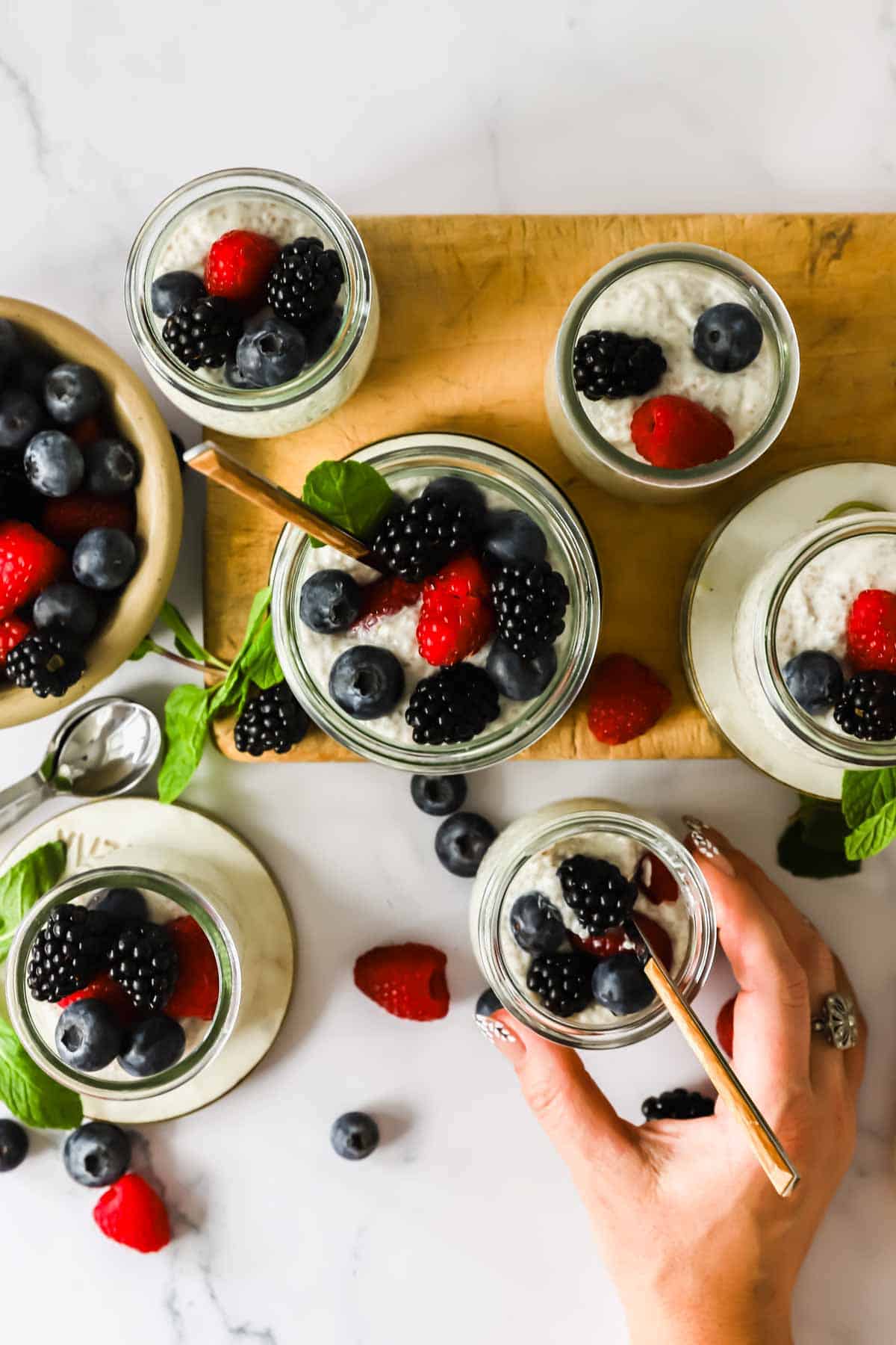 Jars of chia pudding topped with blackberries, blueberries, and raspberries are arranged on a wooden board with a hand reaching for one; extra berries are scattered nearby.