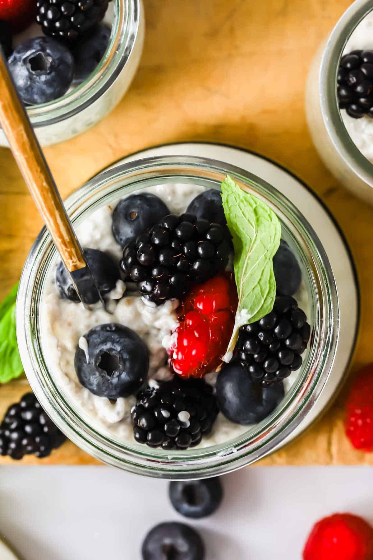 A glass jar filled with overnight chia seed pudding, topped with blueberries, blackberries, a raspberry, and a sprig of mint, with a spoon placed inside.