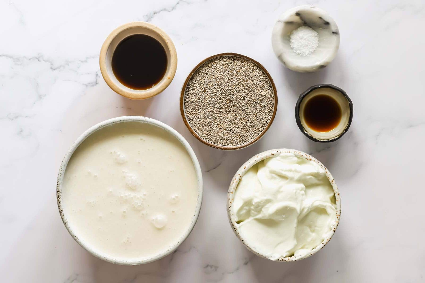 Six bowls on a marble surface containing milk, yogurt, chia seeds, vanilla extract, maple syrup, and salt.