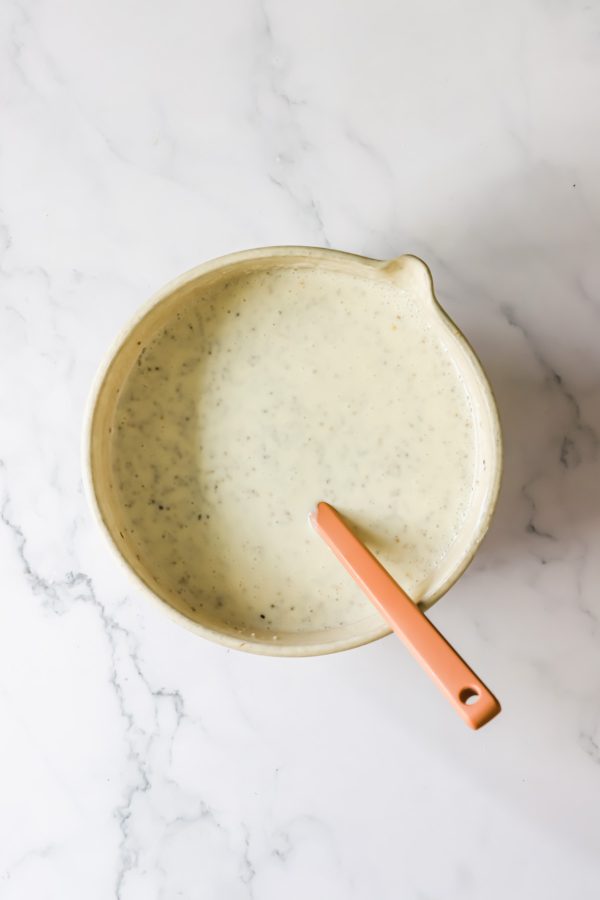 A beige bowl filled with a creamy, speckled chia seed pudding, with an orange-handled spatula resting inside, on a white marble surface.
