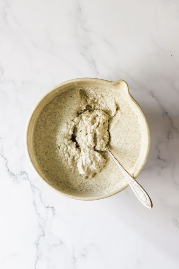 A ceramic bowl of thick, creamy overnight chia seed pudding with a spoon in it, placed on a white marble surface.