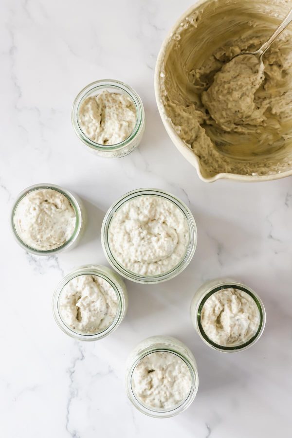 Six glass jars filled with thick, creamy chia seed pudding are arranged on a white surface next to a mixing bowl containing more batter and a spoon.