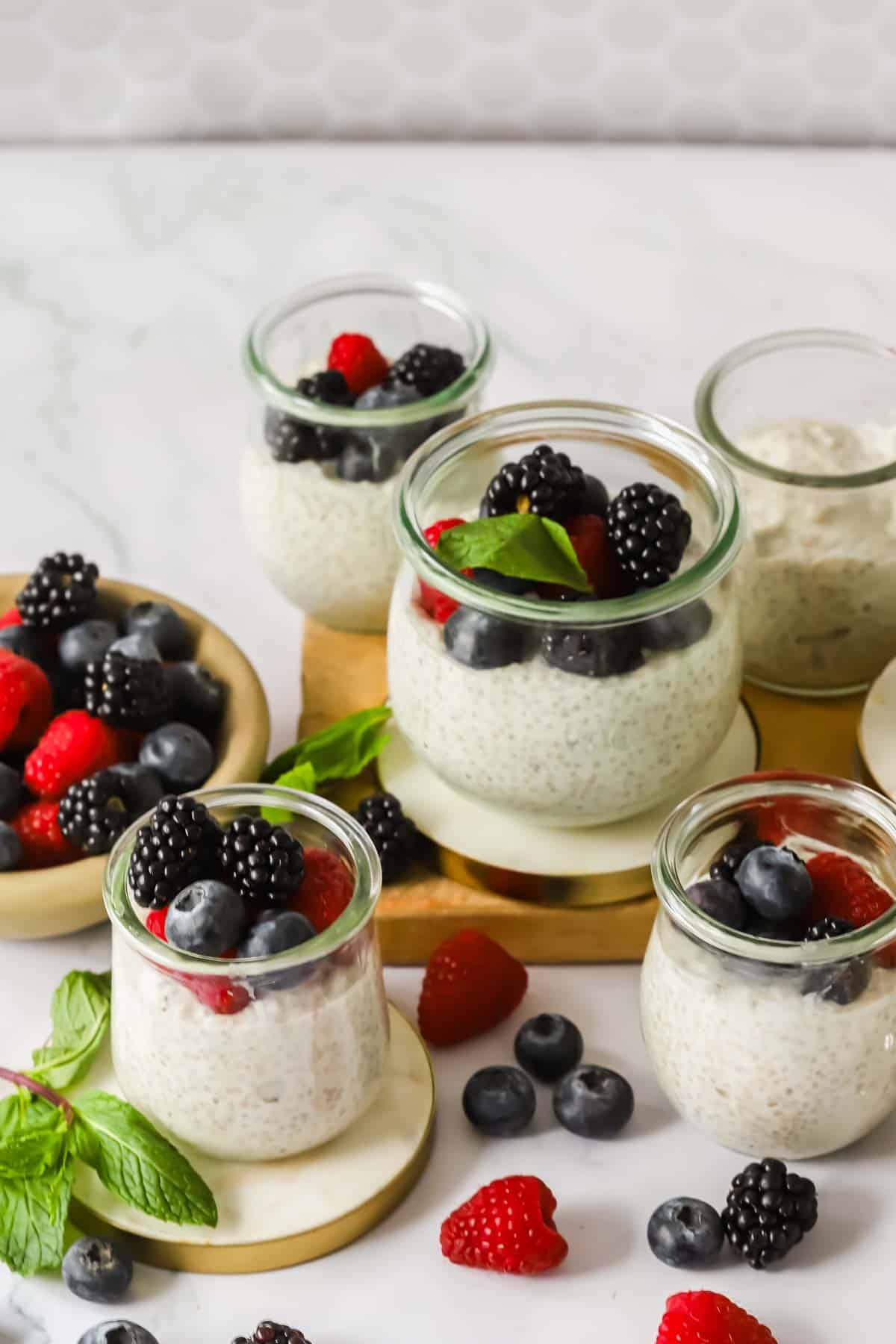 Small glass jars filled with chia seed pudding, topped with blackberries, blueberries, raspberries, and mint leaves, arranged on a white surface with scattered berries and mint.