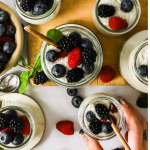 Jars of overnight chia seed pudding topped with blackberries, blueberries, and raspberries are arranged on a table with scattered berries and a hand holding one jar.