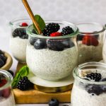 Glass jars filled with chia pudding topped with blackberries, blueberries, and a raspberry, with a gold spoon and a mint leaf for garnish.