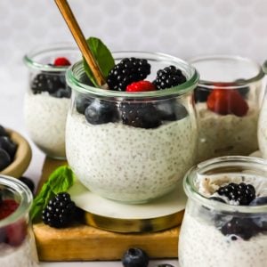 Glass jars filled with chia pudding topped with blackberries, blueberries, and a raspberry, with a gold spoon and a mint leaf for garnish.