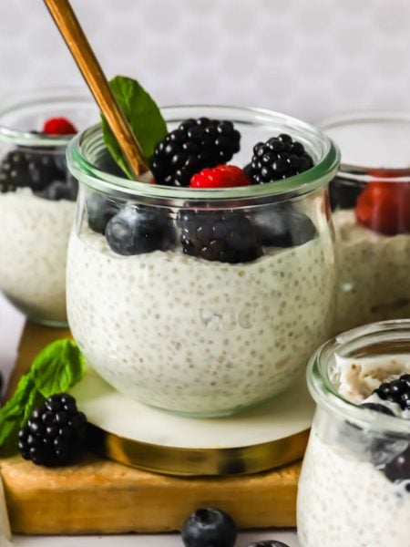 Glass jars filled with chia pudding topped with blackberries, blueberries, and a raspberry, with a gold spoon and a mint leaf for garnish.