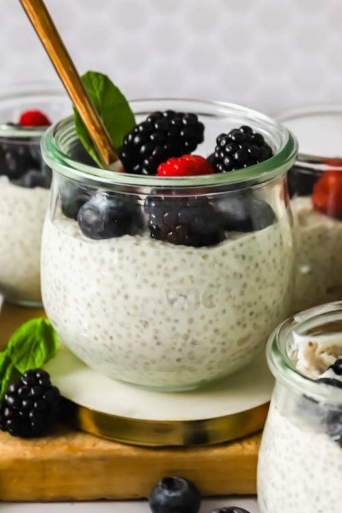 Glass jars filled with chia pudding topped with blackberries, blueberries, and a raspberry, with a gold spoon and a mint leaf for garnish.