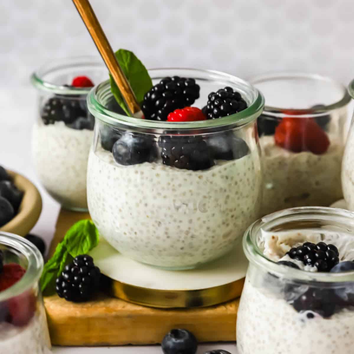 Glass jars filled with chia pudding topped with blackberries, blueberries, and a raspberry, with a gold spoon and a mint leaf for garnish.