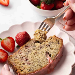 A slice of strawberry banana bread on a plate with fresh strawberries, a fork taking a bite, and lenaskitchenblog.com at the bottom.
