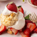 A plate of strawberry shortcake topped with whipped cream and a halved strawberry, with more sliced strawberries on the side and a fork nearby.