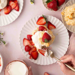 Top-down view of plates with strawberry shortcake, fresh strawberries, whipped cream, and a hand holding a fork and a glass of milk. Text reads: “Easy Easter Dessert Strawberry Shortcake.”.