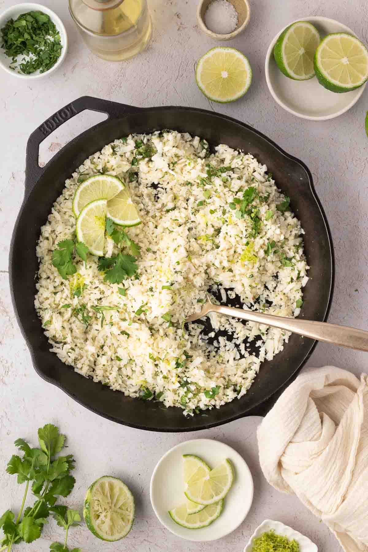 A cast iron skillet filled with cauliflower rice garnished with lime slices and cilantro, surrounded by fresh lime, cilantro, and a glass of white wine.