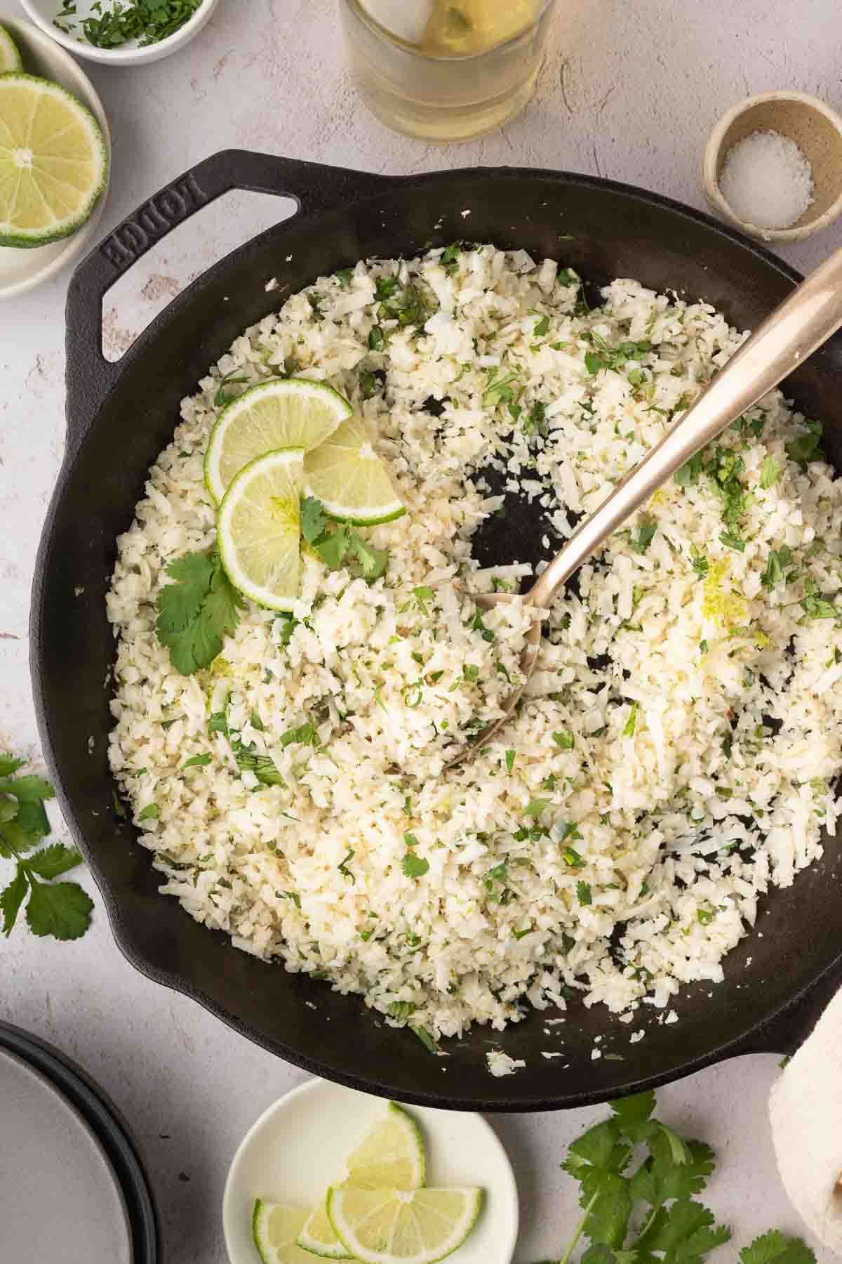 A black skillet filled with cauliflower rice garnished with lime slices and cilantro, with a serving spoon resting inside. Small bowls of ingredients surround the skillet.