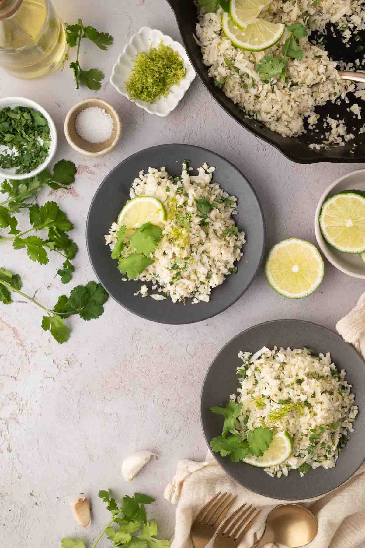 Two plates of cauliflower rice garnished with lime slices and cilantro are on a table, surrounded by fresh herbs, lime, seasonings, and a skillet with more cauliflower rice.