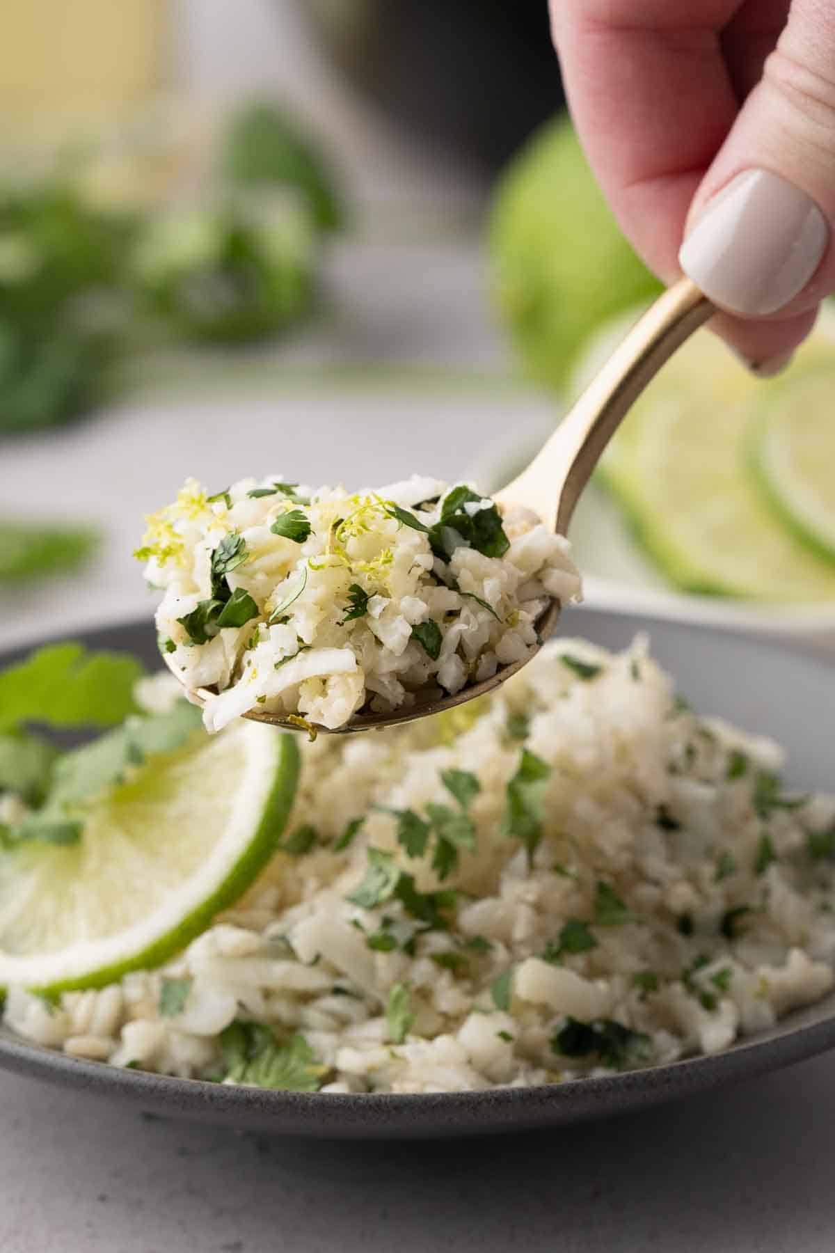 A spoonful of cauliflower rice garnished with herbs, held above a plate with more cauliflower rice and a slice of lime.