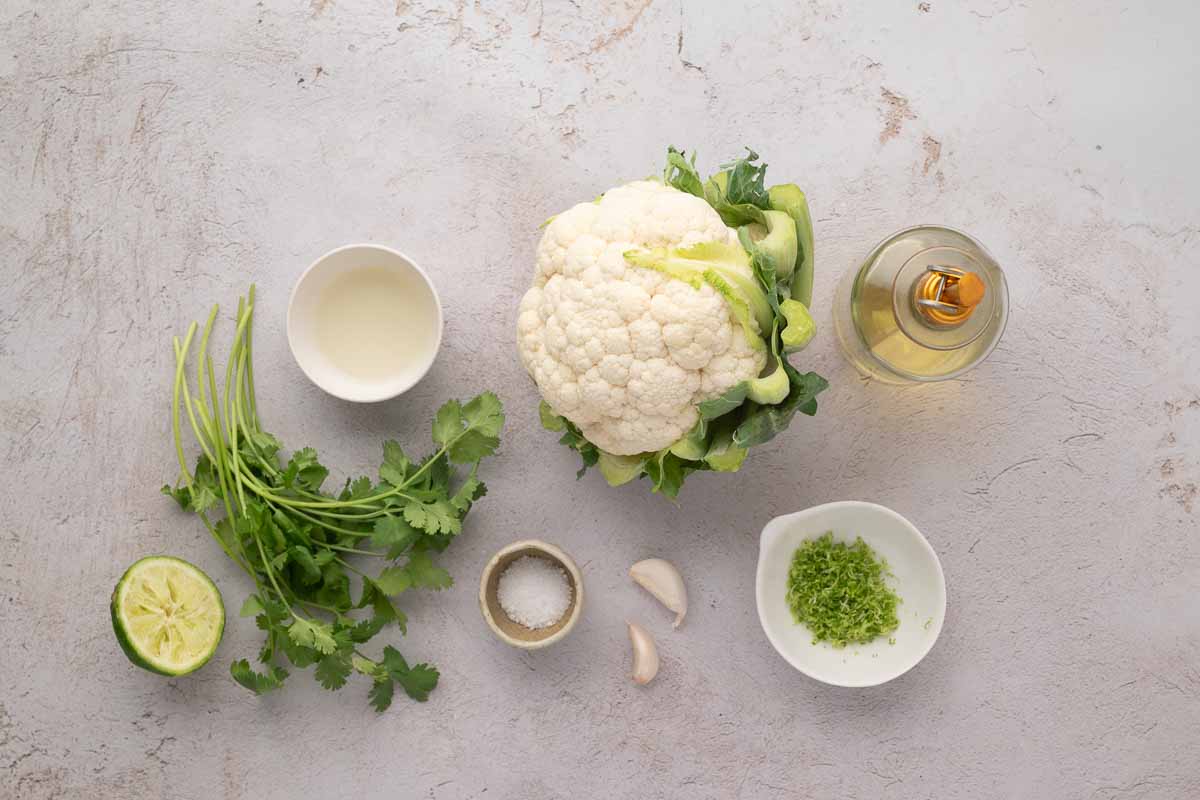 A head of cauliflower, fresh cilantro, half a lime, a small bowl of cream, a bowl of oil, a bowl of salt, two garlic cloves, and a bowl of lime zest arranged on a light surface.