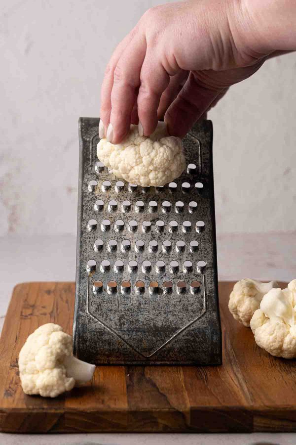 A hand holding a cauliflower floret over a metal grater on a wooden board, with additional cauliflower pieces nearby.