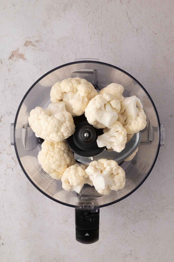 Raw cauliflower florets placed inside a food processor, viewed from above on a light-colored surface.