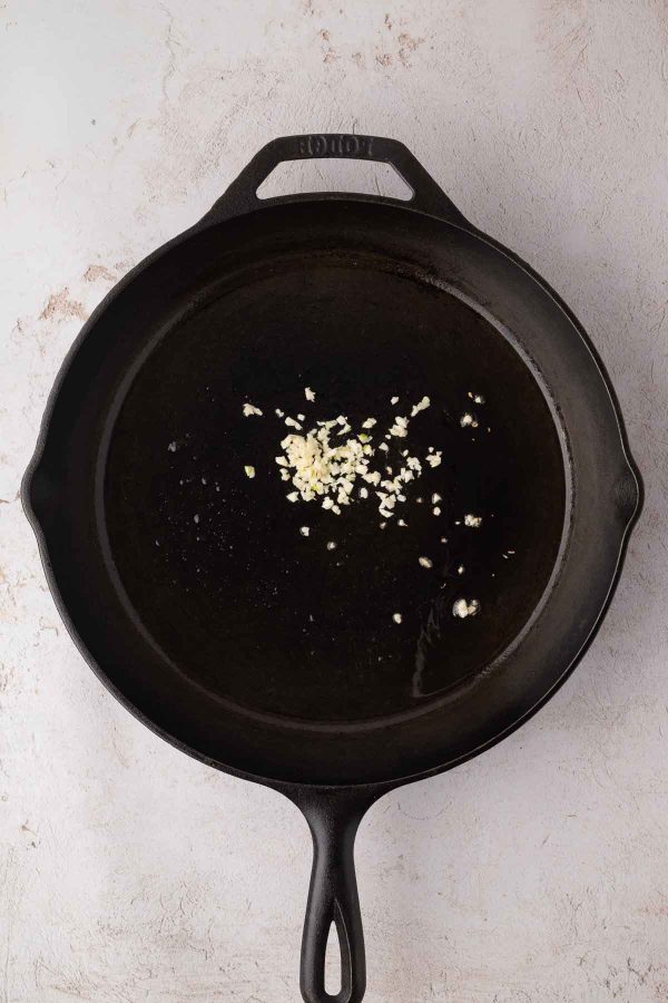 A black cast iron skillet with a small amount of minced garlic and oil inside, placed on a light-colored surface.