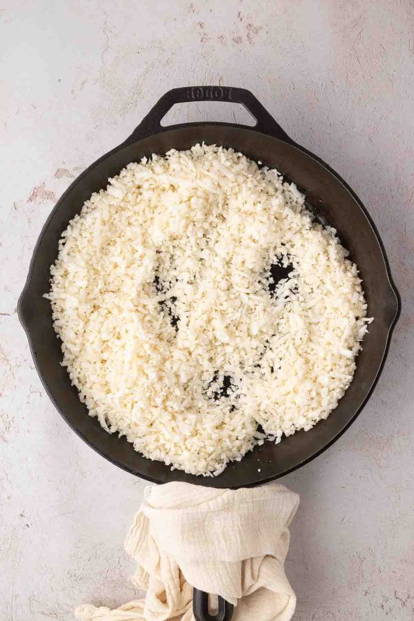 A cast iron skillet filled with cooked white rice sits on a light-colored surface, with a beige towel wrapped around the handle.