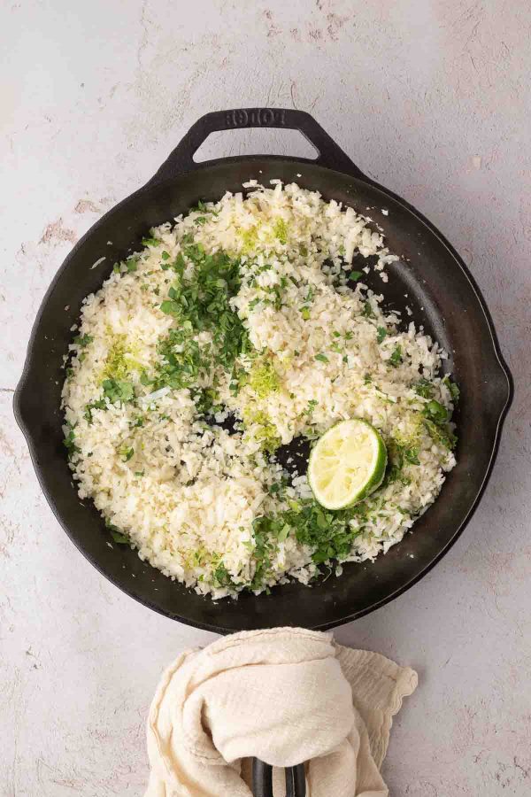 A cast iron skillet filled with cooked rice garnished with chopped herbs and half a lime, placed on a light surface with a beige towel nearby.