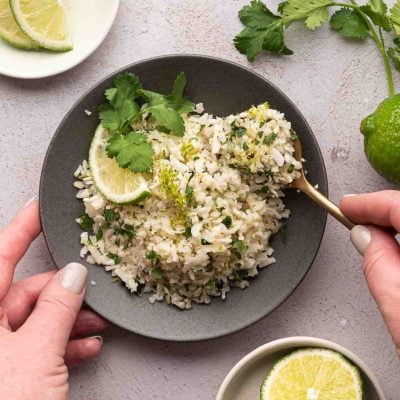 A bowl of cilantro lime cauliflower rice garnished with chopped herbs, a lime wedge, and cilantro, with a hand holding a spoon and another hand holding the bowl. Lime slices and cilantro are nearby.