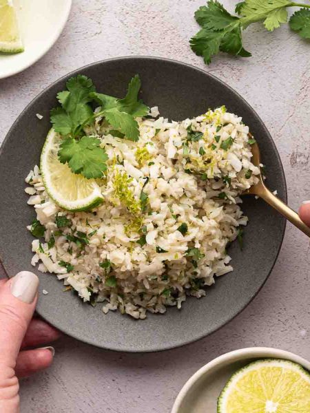A bowl of cilantro lime cauliflower rice garnished with chopped herbs, a lime wedge, and cilantro, with a hand holding a spoon and another hand holding the bowl. Lime slices and cilantro are nearby.