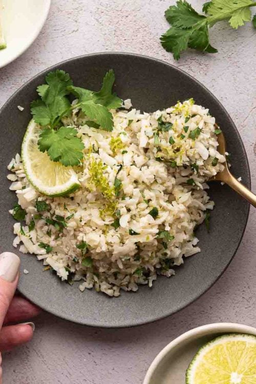 A bowl of cilantro lime cauliflower rice garnished with chopped herbs, a lime wedge, and cilantro, with a hand holding a spoon and another hand holding the bowl. Lime slices and cilantro are nearby.
