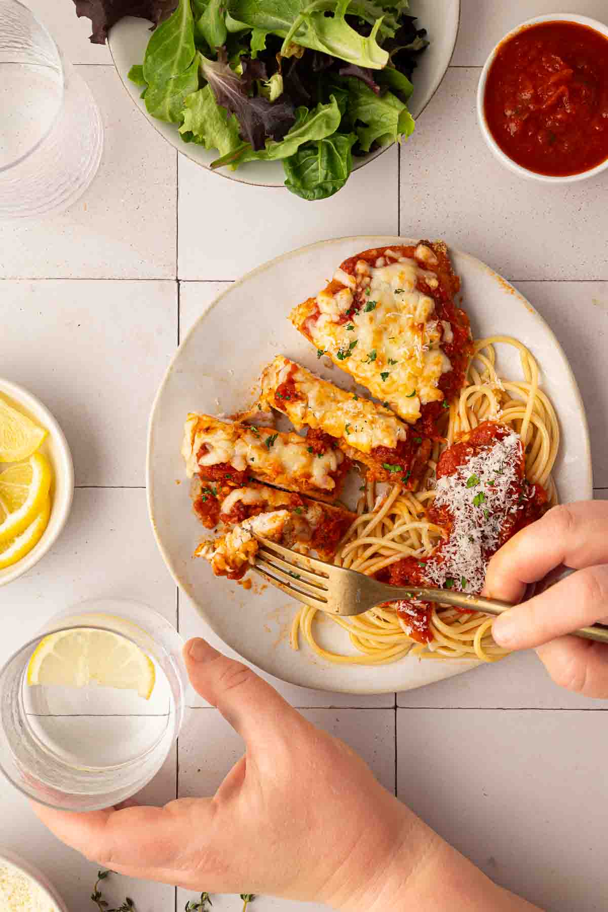 A plate of sheet pan chicken parmesan with spaghetti and tomato sauce, a side salad, lemon slices, and a hand holding a fork and a glass of water.